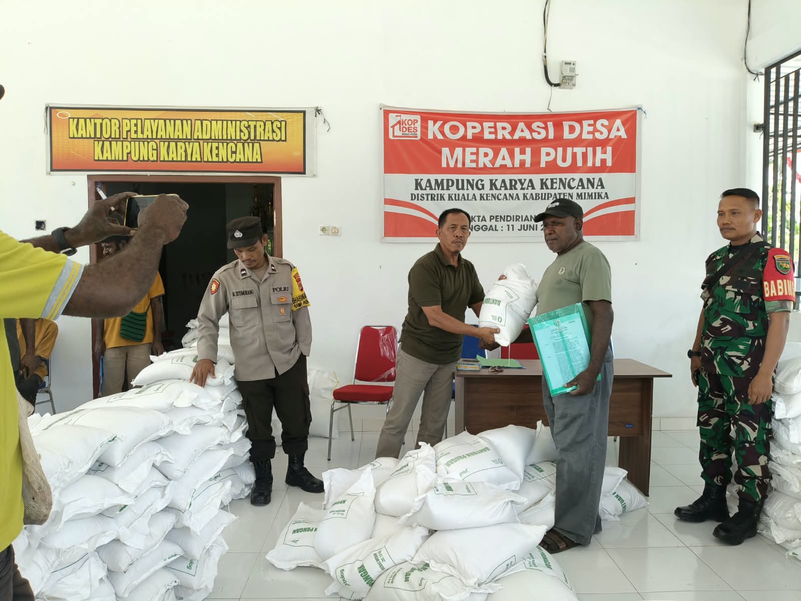 Kepala Kampung Karya Kencana, Rakam Suntoro menyerahkan bantuan beras kepada warga disaksikan oleh Babinsa Koramil Kuala Kencana, Bhabinkamtibmas Kampung Karya Kencana dan warga yang hadir 